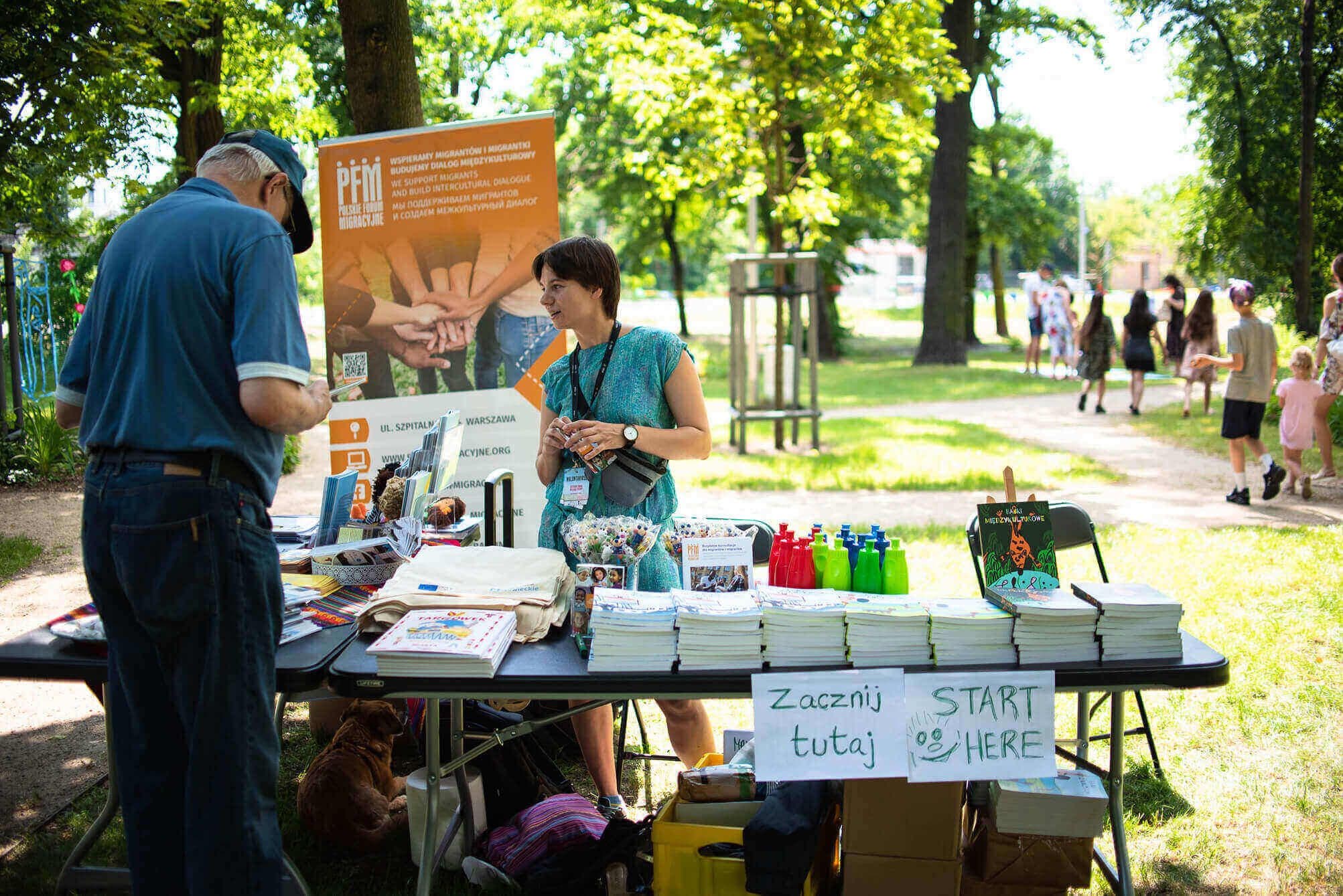 Wolontariuszka przy stoisku z materiałami informacyjnymi organizacji migranckiej w parku