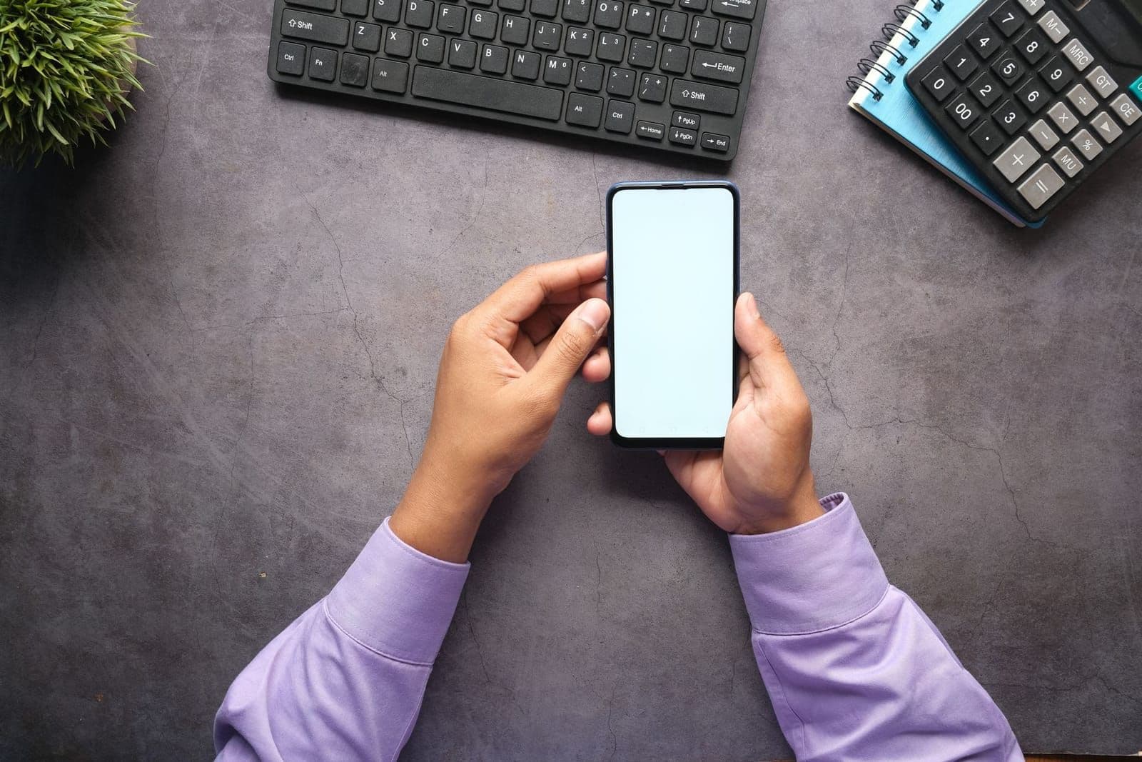 Dłonie trzymają smartfon nad biurkiem z klawiaturą